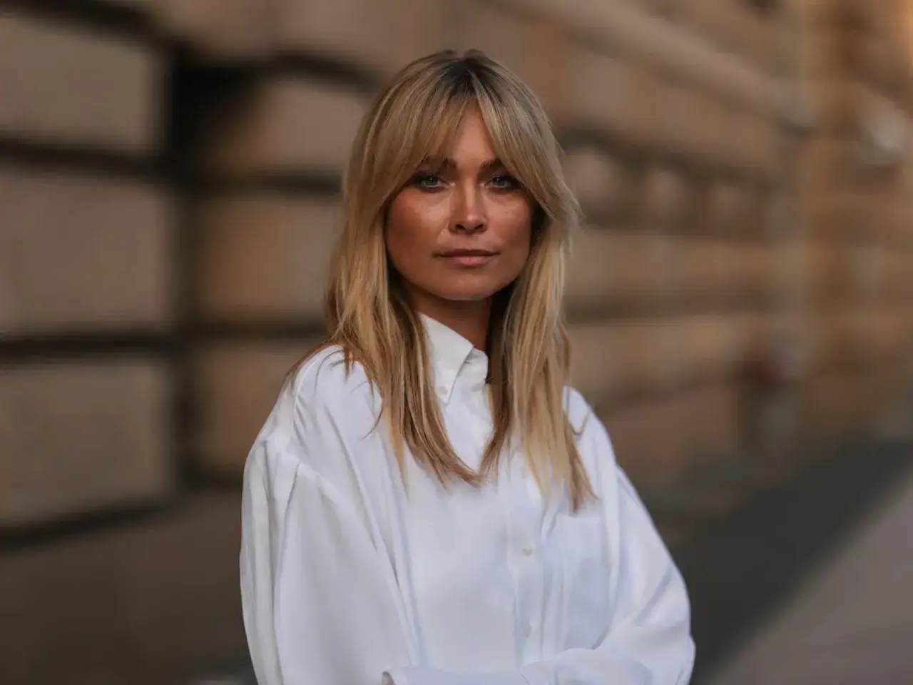 Frau mit blonden Haaren und **wispy bangs**, trägt ein weißes Hemd.