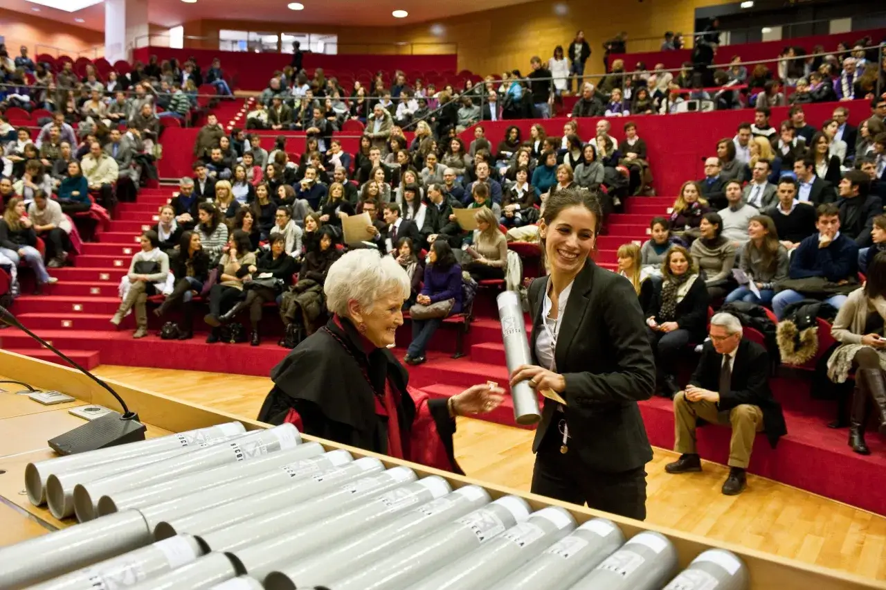 Una giovane laureata magistrale sorride mentre riceve il suo diploma in un'aula gremita di studenti e docenti.