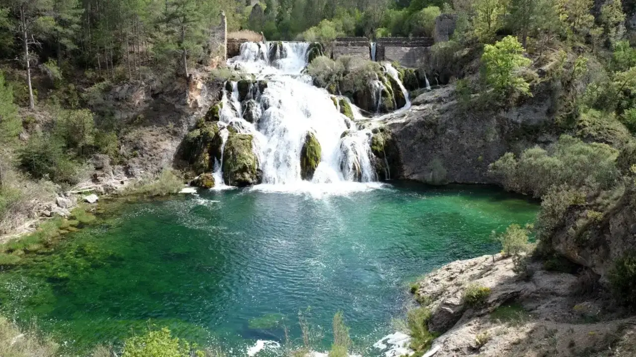 Cascada de agua blanca cayendo en **piscinas naturales** de color verde esmeralda, rodeadas de vegetación y rocas.
