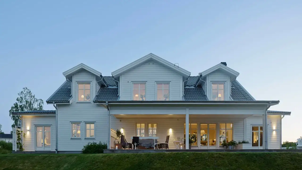 Vackert vitt hus med veranda, byggt i gammal stil. Ljuset fr&aring;n f&ouml;nstren skapar en varm och inbjudande atmosf&auml;r.