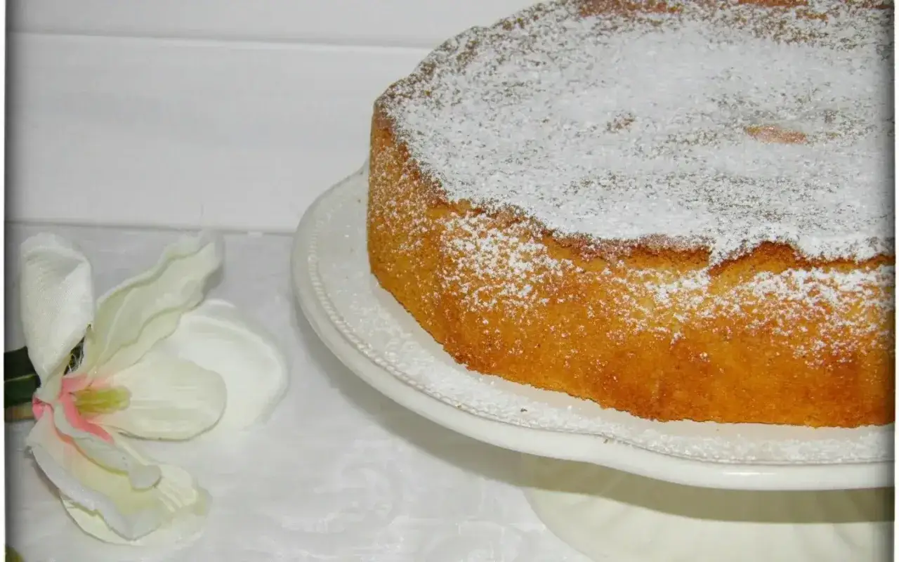 Ein goldbrauner mallorquinischer Zitronenkuchen, bestäubt mit Puderzucker, neben einer weißen Blüte.