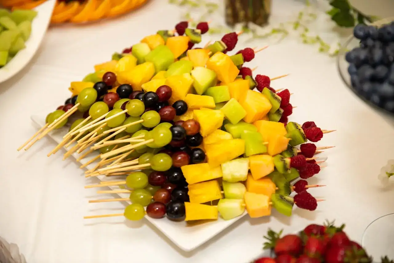 Kolorowe szaszłyki z owoców: winogrona, kiwi, mango, melony i maliny na białym talerzu.