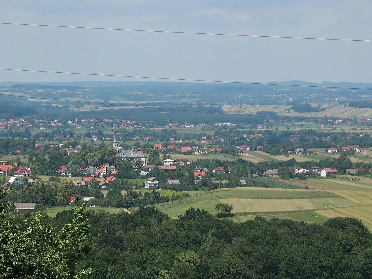 Widok na rozległą wieś Przytkowice z kościołem w centrum, otoczoną zielonymi polami i lasami.