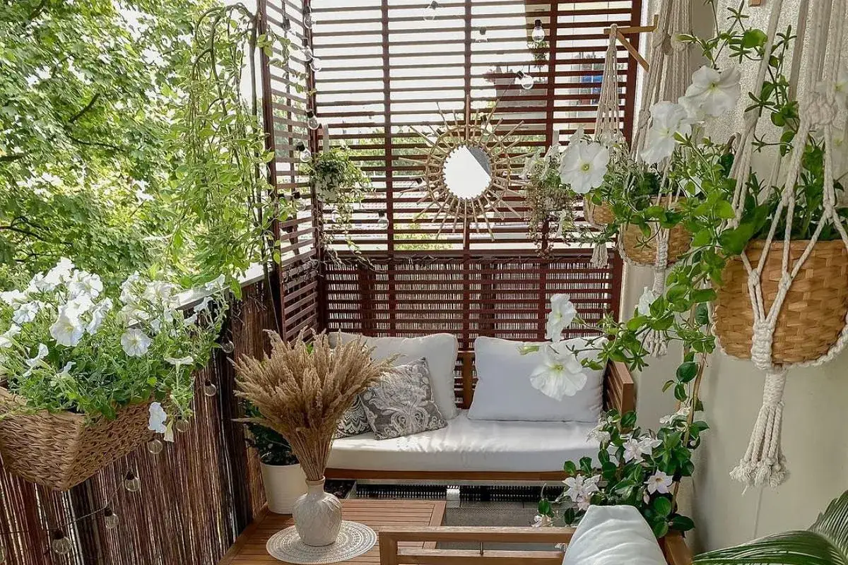 Przytulny balkon w bloku, jak osłonić go roślinnością i drewnianymi panelami. Białe kwiaty, makramy i słoneczne lustro tworzą relaksującą atmosferę.