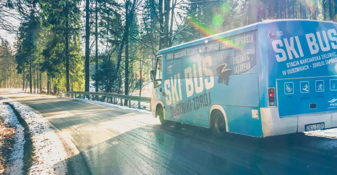 Ski Bus w Dusznikach-Zdroju zaprasza na stoki narciarskie w Zieleńcu. Autobus jedzie zimową drogą wśród drzew.