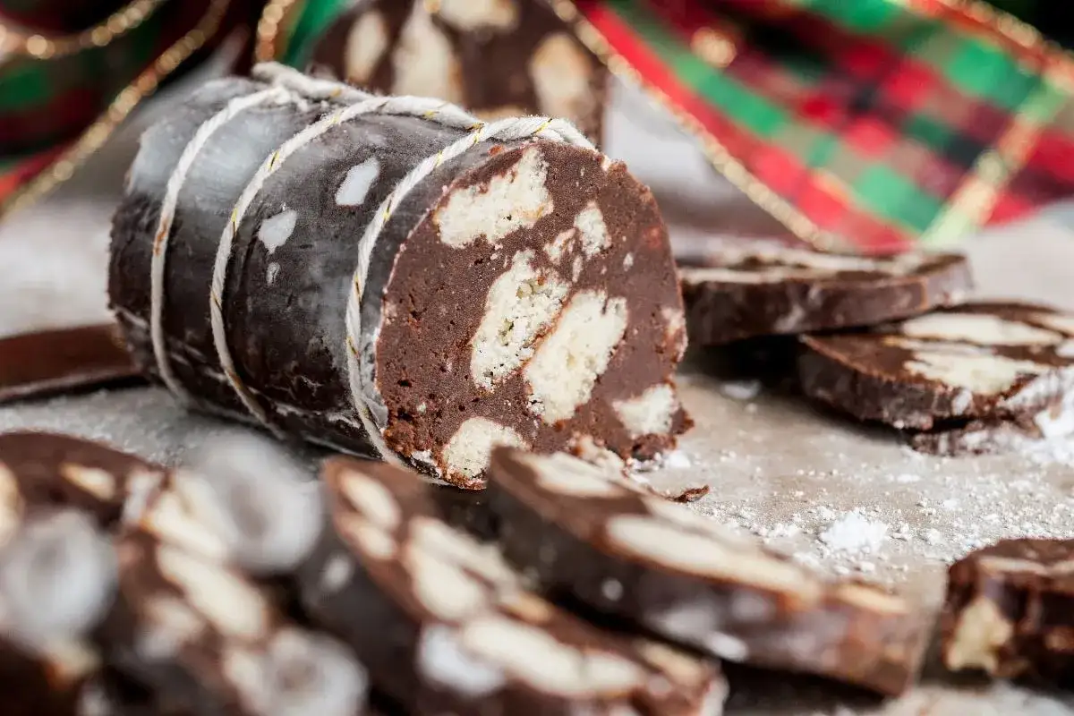 Ein köstlicher salame al cioccolato, gebunden mit Schnur, mit Scheiben daneben und weihnachtlichem Band im Hintergrund.