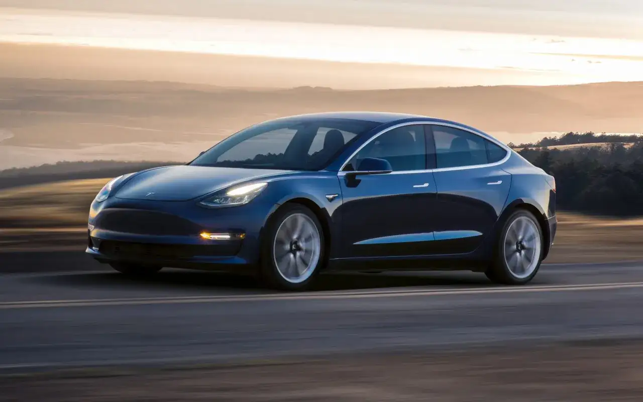 Niebieska Tesla pędzi po drodze, osiągając prędkość do 100 km/h. W tle zachodzące słońce i krajobraz.