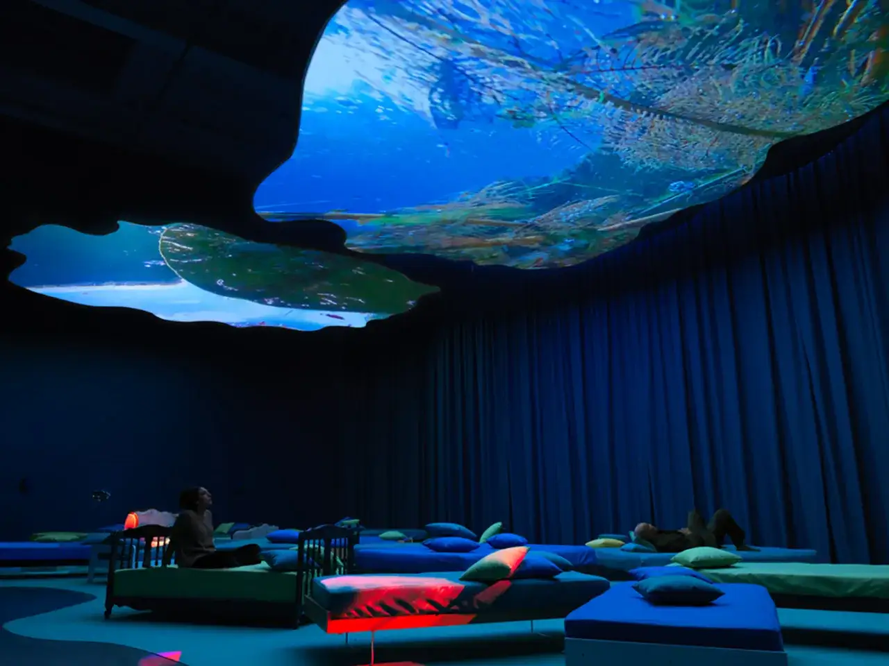 An immersive gallery exhibition features beds arranged in a dark room, with a projected underwater scene on the ceiling, creating a dreamlike experience.