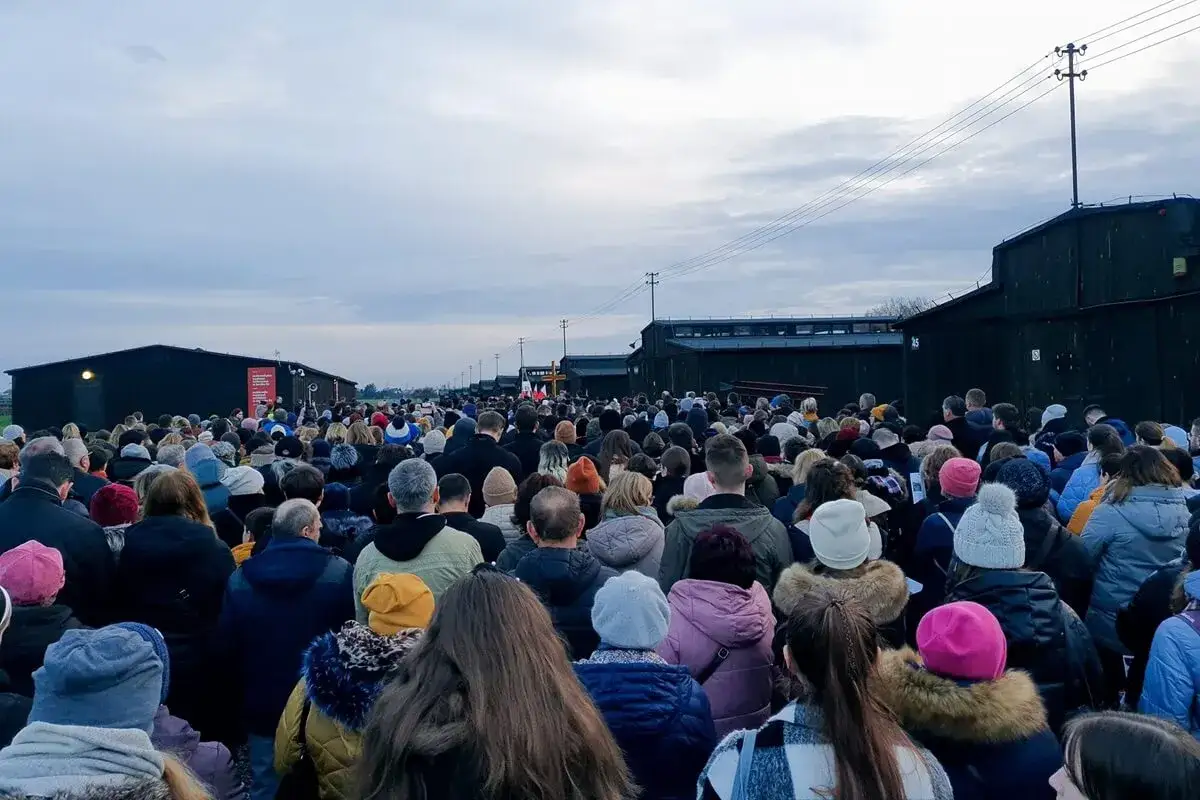 Tłum ludzi uczestniczy w Drodze Krzyżowej na Majdanku, stojąc przed ciemnymi barakami.