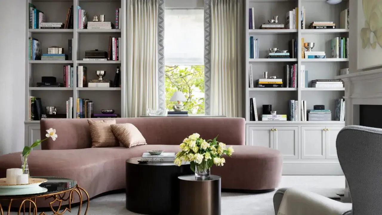 Soggiorno elegante con divano curvo rosa cipria, librerie piene di libri e oggetti, tavolini moderni e un vaso di fiori gialli. Idee originali per arredare soggiorno.