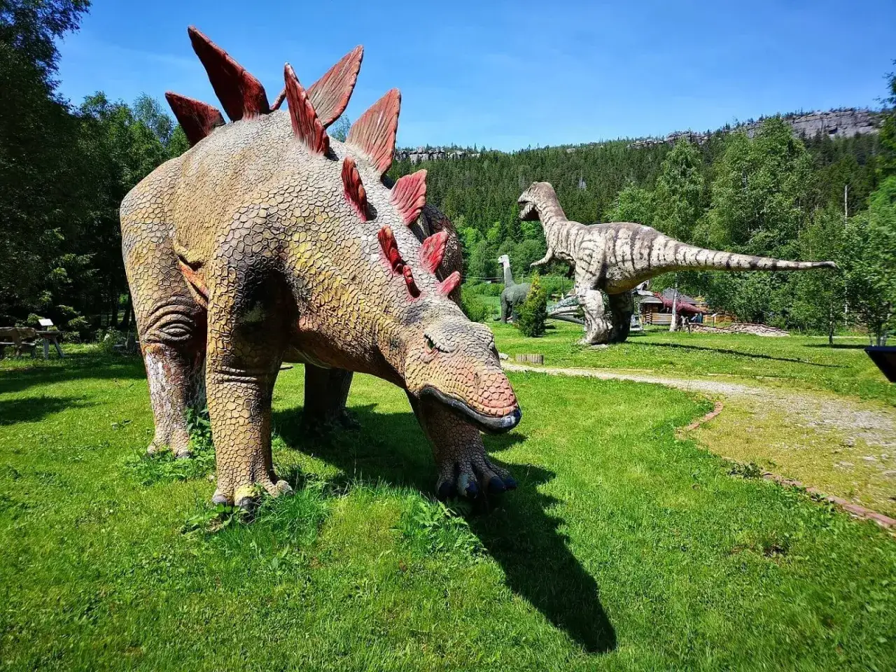 W parku dinozaurów karłów Stegozaur pochyla się do trawy, podczas gdy Tyranozaur i inne prehistoryczne stworzenia stoją w oddali na tle zielonych drzew.