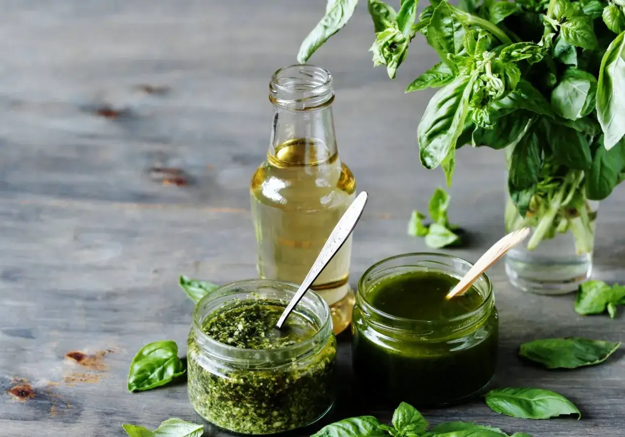 Dwa słoiczki z zielonym pesto i butelka oliwy, obok bukiet świeżej bazylii. Idealne składniki na orzeźwiający drink z bazylia.