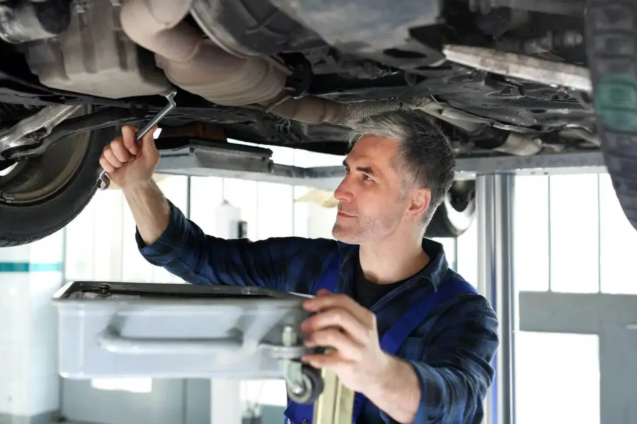 Mechanik dokonuje wymiany miski olejowej, używając klucza do odkręcenia śrub. Pod samochodem stoi pojemnik na zużyty olej.