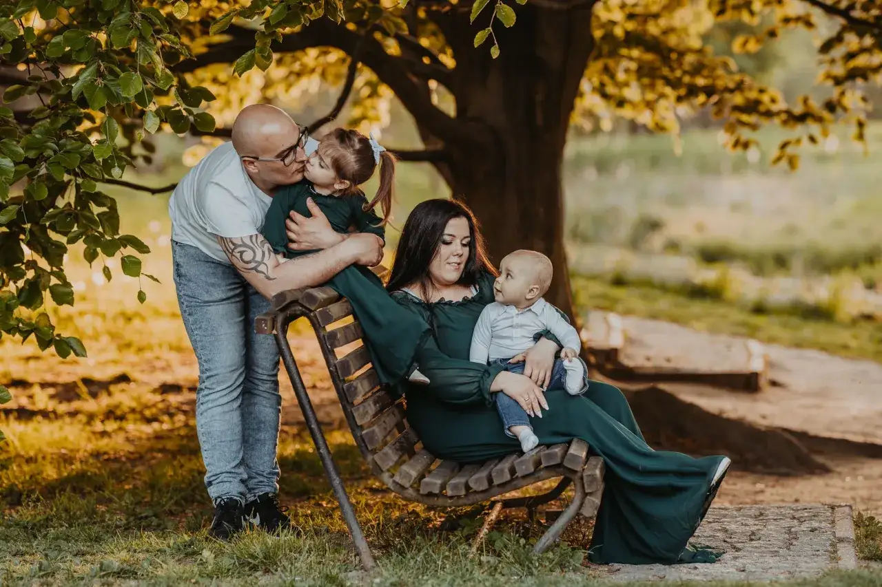 Rodzina na ławce w parku. Ojciec całuje córkę, matka trzyma niemowlę. Piękna fotografia plenerowa w złotym świetle.