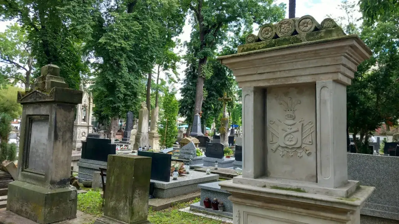 Kamienny nagrobek z herbem, ozdobiony motywami roślinnymi, stanowi historyczne epitafium. Wokół wiele innych nagrobków i zieleni.