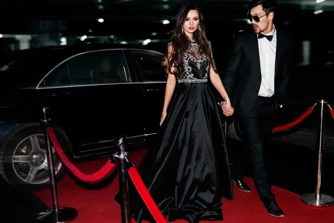 Pareja con vestir formal camina por alfombra roja junto a coche de lujo.