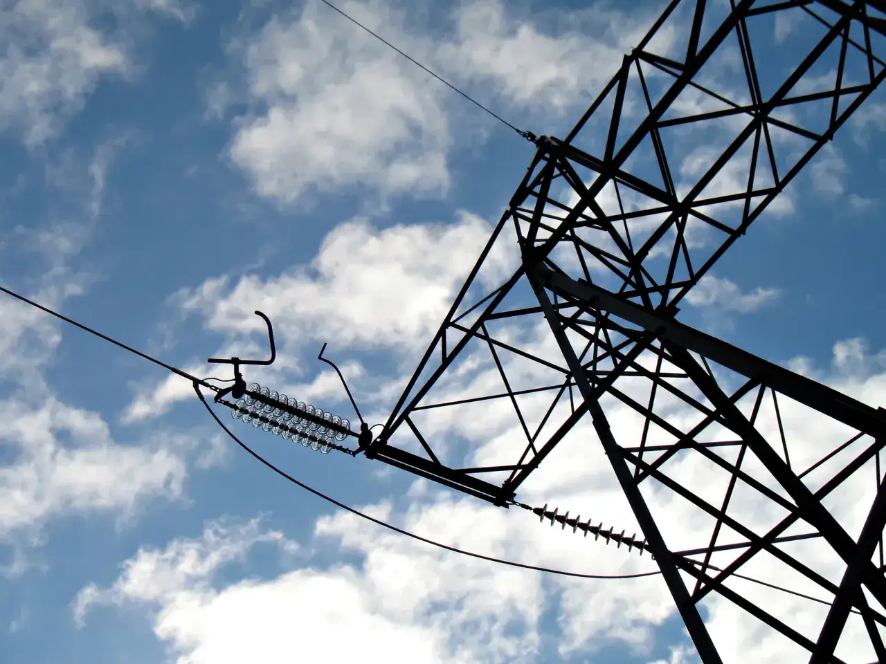 Siatka stalowej konstrukcji wieży energetycznej na tle błękitnego nieba z chmurami. Widoczne izolatory i przewody, przypominające o konieczności zachowania bezpieczna odległość od linii wysokiego napięcia 110 kv.