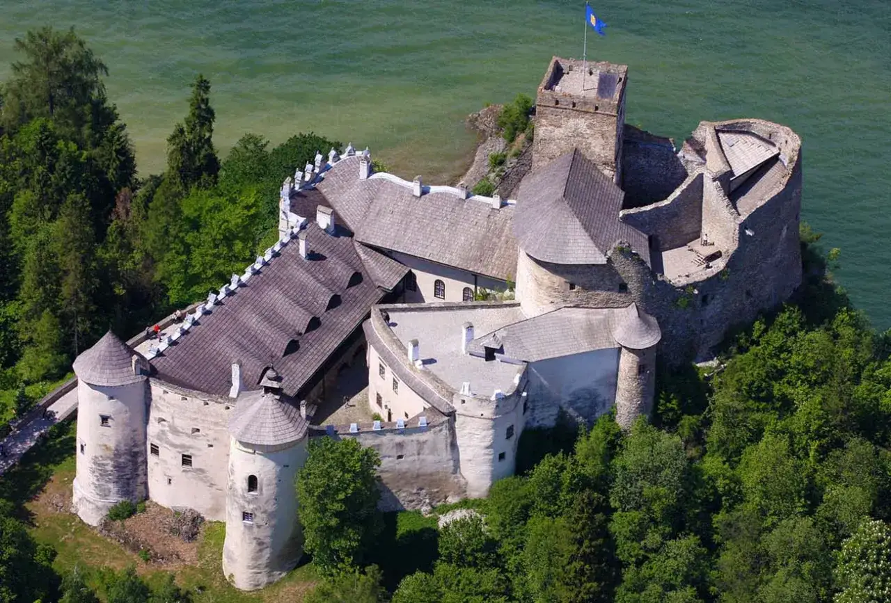 Zamek w Niedzicy, malowniczo położony nad jeziorem, z wieżą i murami obronnymi, otoczony zielenią.