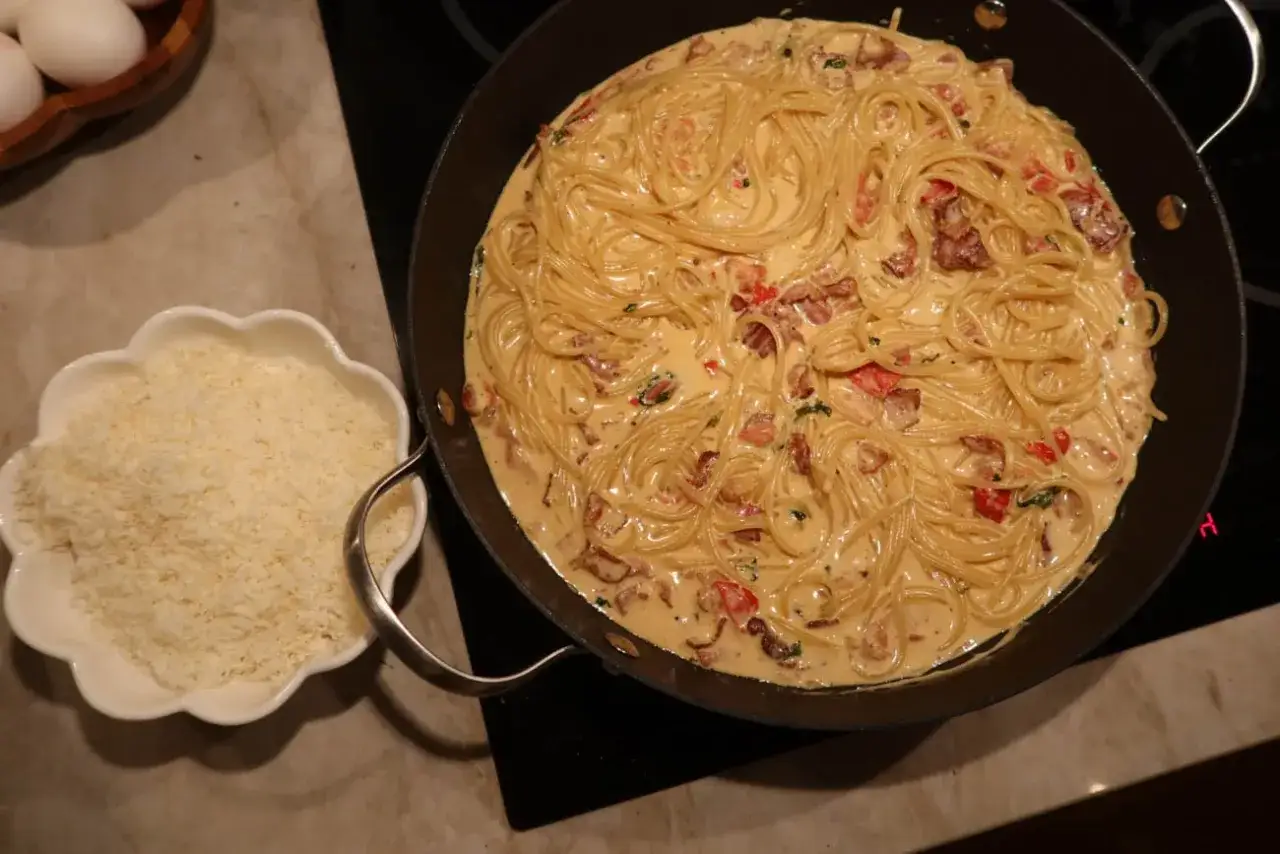 En stor panna med krämig pasta med baconsås, bredvid en skål med riven ost.