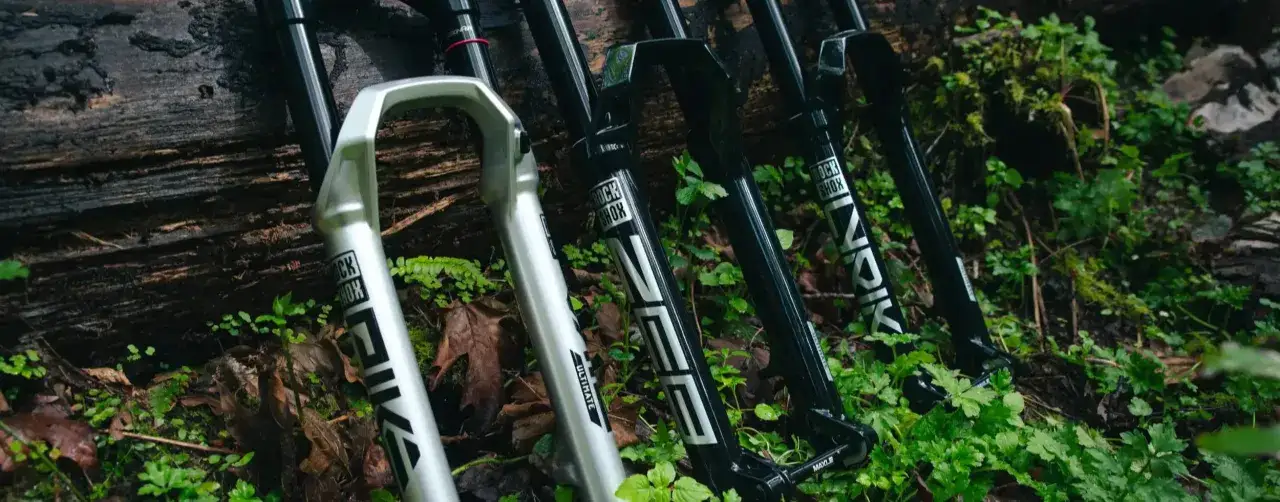 A silver RockShox Pike and black RockShox Lyrik forks lean against a log, ready for trail adventures.