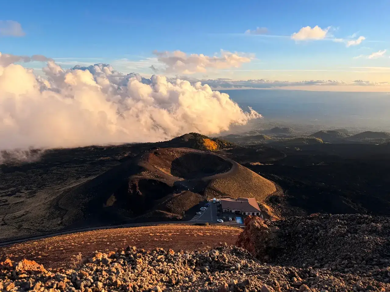 Un paesaggio lunare con un cratere e un rifugio, nuvole dorate e il sole che tramonta. Un'esperienza indimenticabile per visitar el Etna.