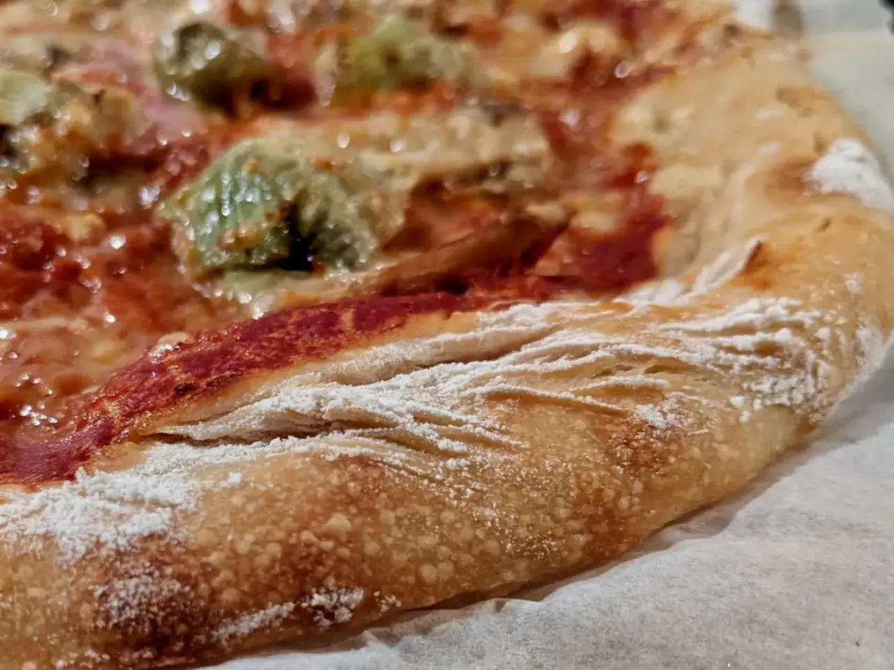 Gros plan sur la cro&ucirc;te dor&eacute;e et a&eacute;r&eacute;e d'une pizza, saupoudr&eacute;e de farine. On aper&ccedil;oit la garniture rouge et verte. Un vrai d&eacute;lice de pain au levain.