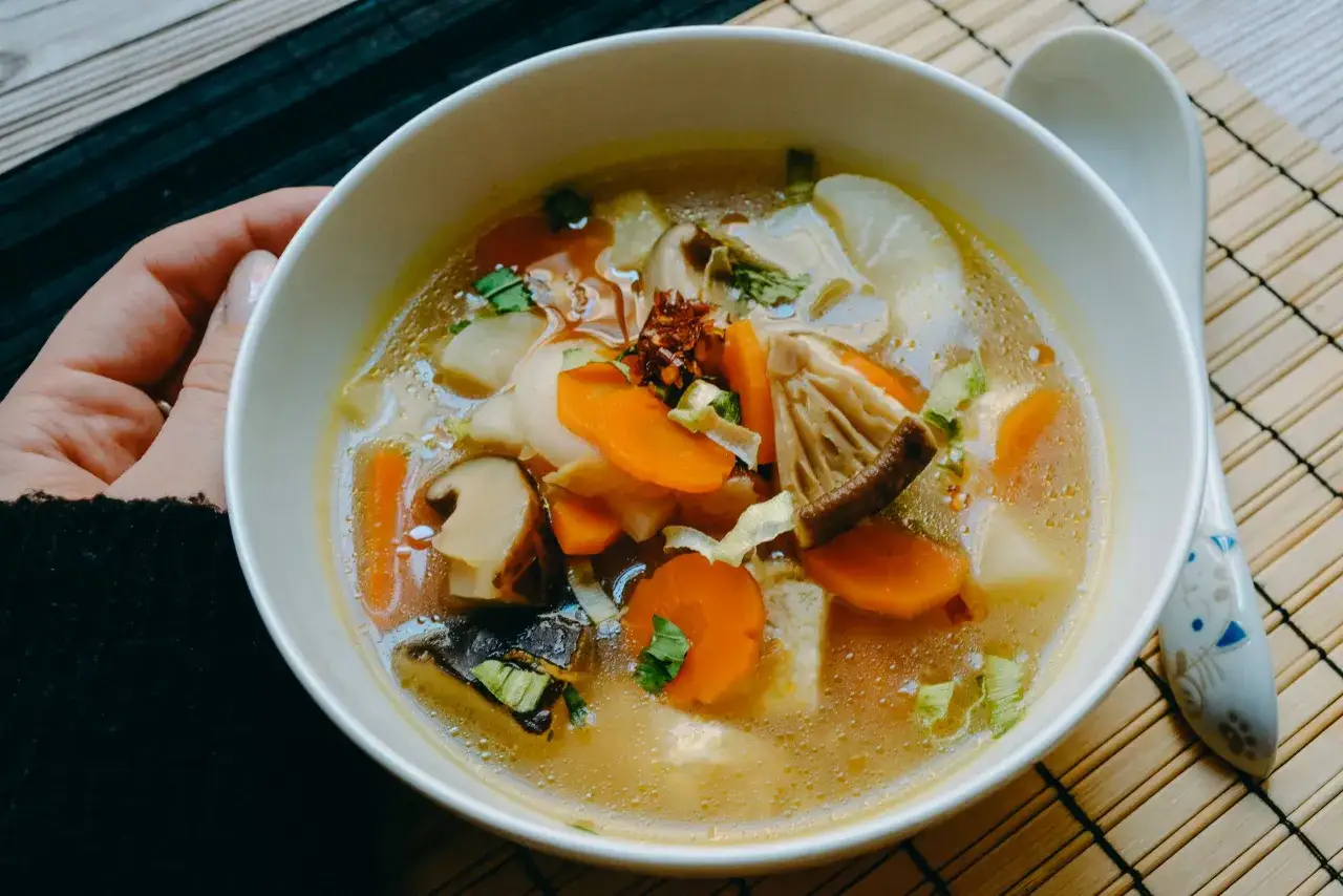 A steaming bowl of Japanese vegetable soup, brimming with carrots, mushrooms, and daikon radish, is held by a hand.