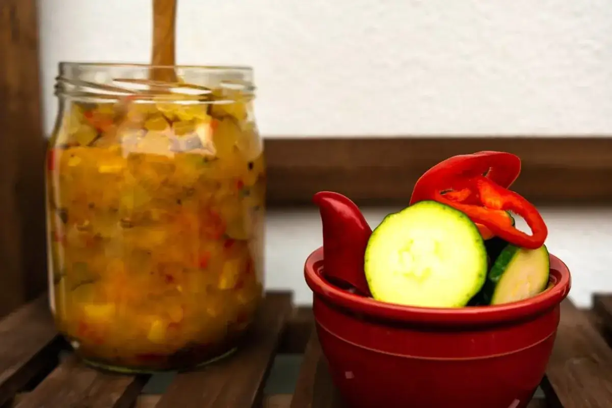 Ein Glas mit würzigem Relish steht neben einer roten Schale mit Zucchini und Paprika.