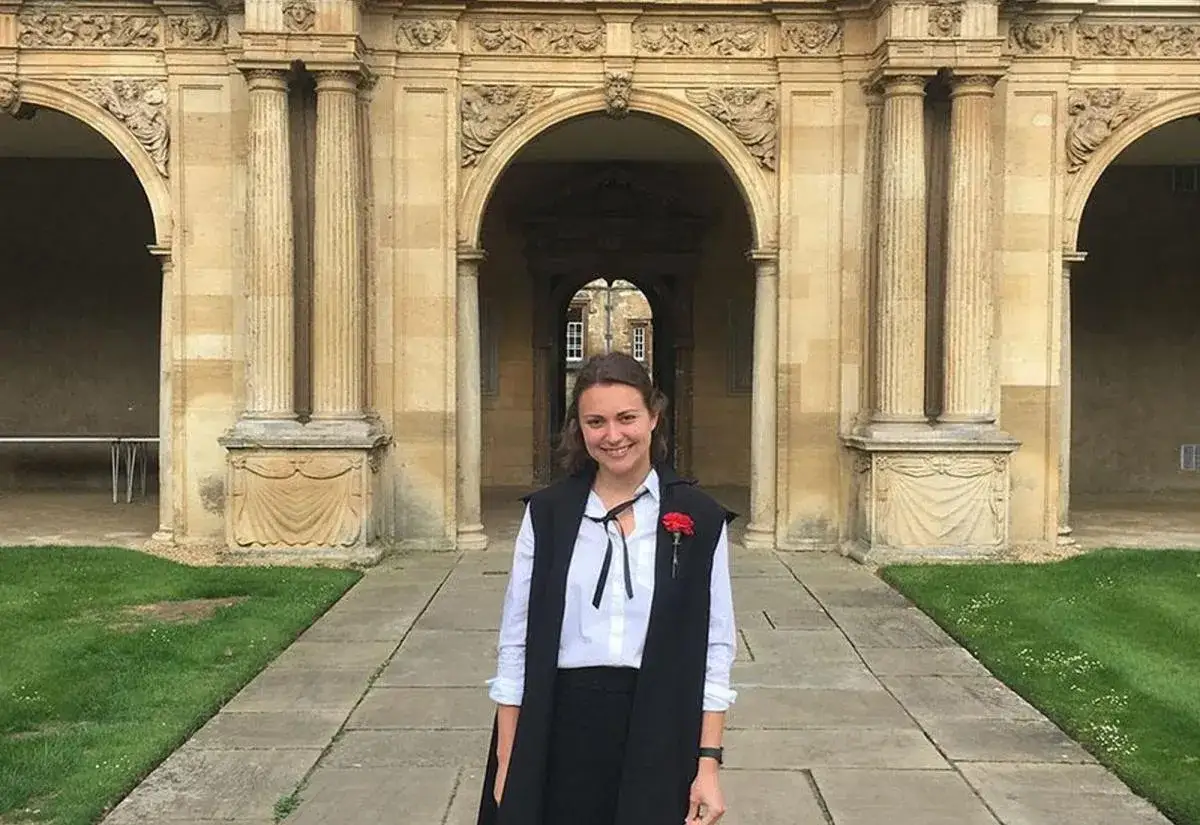 A smiling graduate in academic regalia stands before an ornate stone archway, celebrating her government degree.