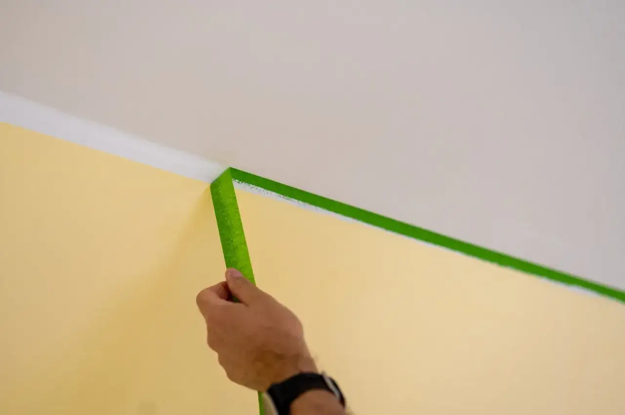 Hand klebt grünes Malerkrepp an der Wand, um die Decke zu streichen.