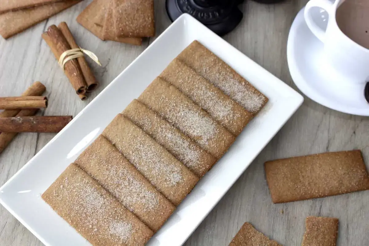 Galletas rectangulares espolvoreadas con azúcar y canela, junto a palitos de canela y una taza de chocolate caliente.