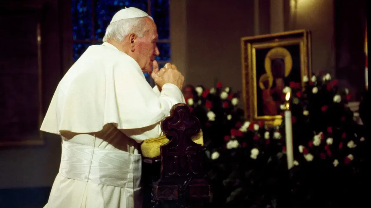 Papież Jan Paweł II w modlitwie. W tle obraz Matki Boskiej i kwiaty.
