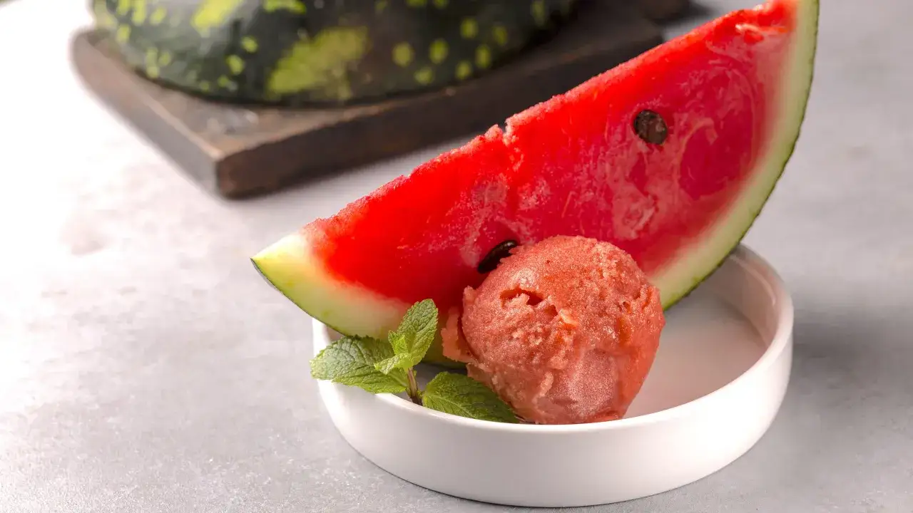 Una pallina di gelato anguria fatto in casa, rinfrescante e profumata, accanto a una fetta di anguria e foglie di menta.