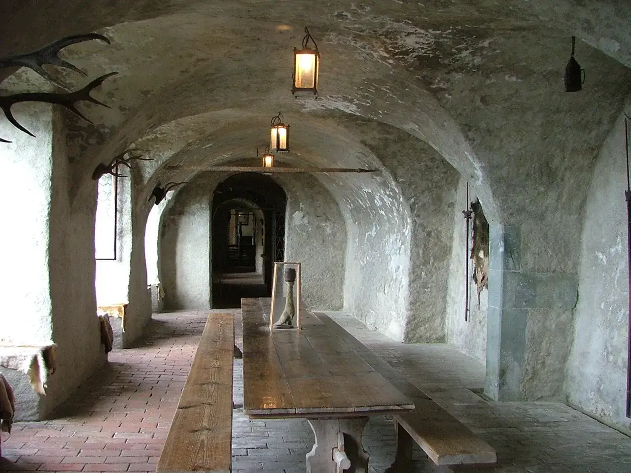 Langer Holztisch mit B&auml;nken in einem Gew&ouml;lbekeller des Meersburg Castle. Hirschgeweihe und alte Laternen schm&uuml;cken die W&auml;nde.