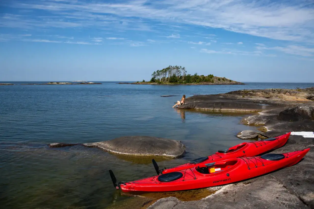 Tv&aring; r&ouml;da kajaker ligger p&aring; en klippig strand. En kvinna sitter p&aring; en sten och blickar ut &ouml;ver vattnet. En perfekt dag f&ouml;r kajak i Karlstad.