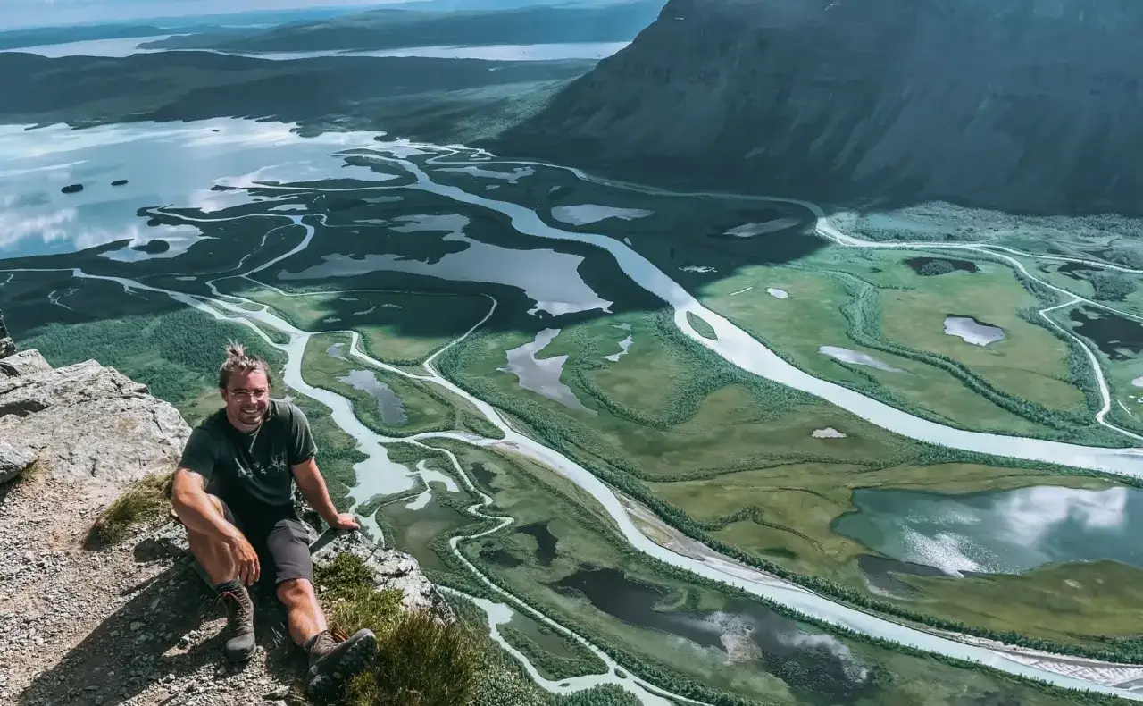 Man sitter p&aring; en klippa med en fantastisk utsikt &ouml;ver ett meandrande vattendrag och fj&auml;llandskap i Sarek. Aktse till skierfe!