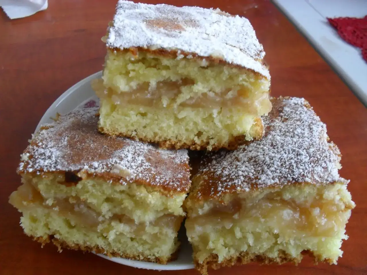 Trzy kawałki najprostszego jabłecznika, posypane cukrem pudrem, leżą na talerzyku.