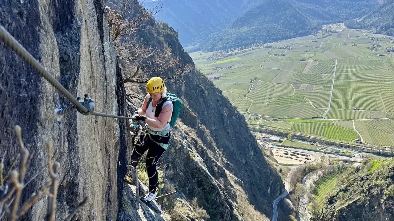 Frau im Helm klettert gesichert am Drahtseil den **Hoachwool Klettersteig** entlang, mit Blick auf Weinberge.