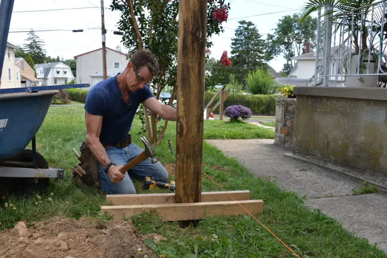 Man demonstrates how to install fence post by hammering wooden braces around a new post.