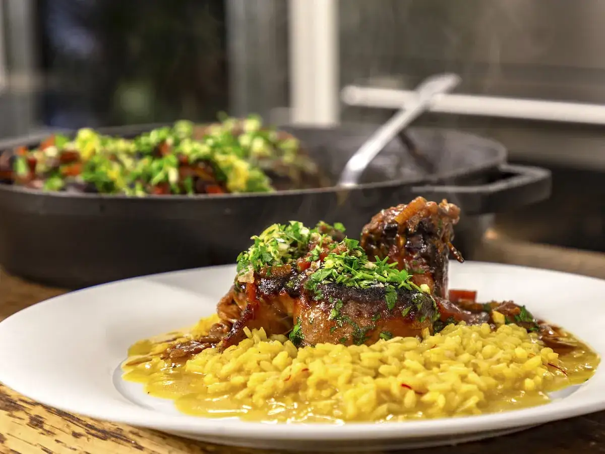 En tallrik med osso buco, saftigt k&ouml;tt med ben, serverat p&aring; en b&auml;dd av gyllene risotto. I bakgrunden syns en gryta med gr&ouml;nsaker.