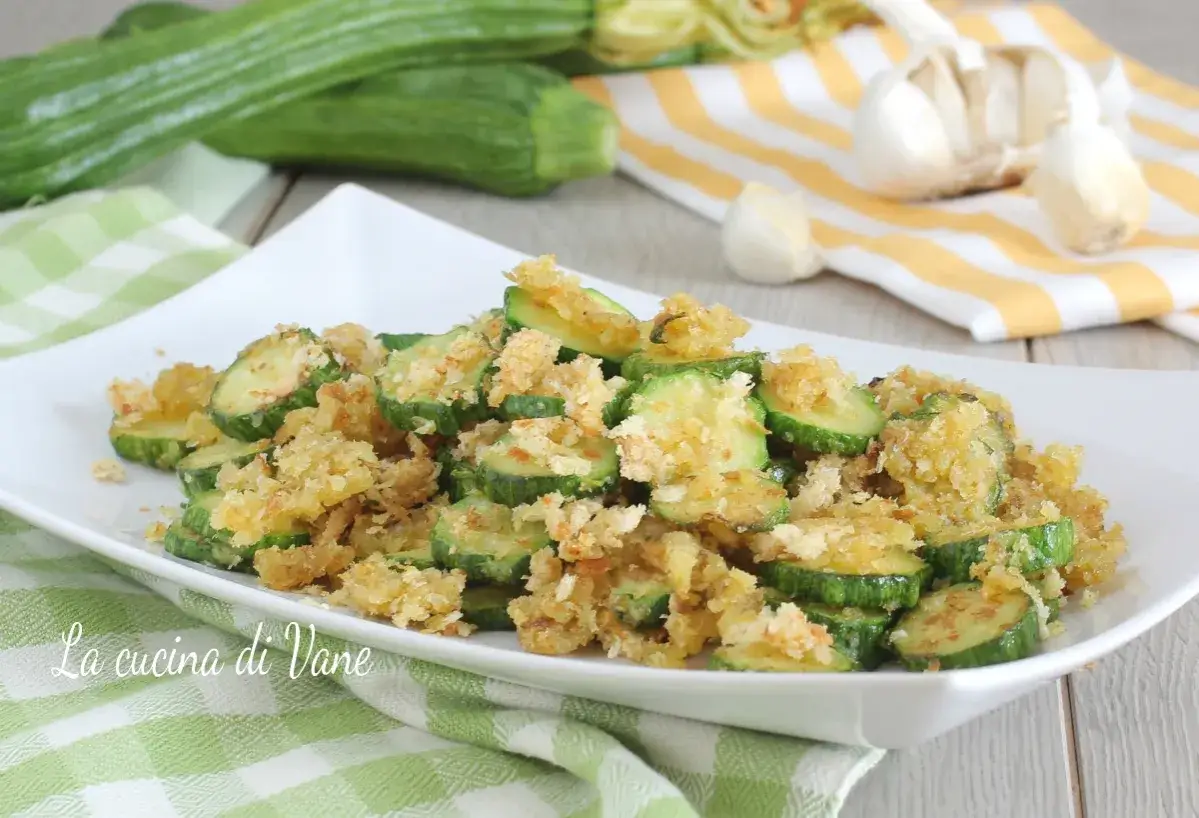 Zucchine sabbiose in padella, croccanti e dorate, servite su un piatto bianco. Sullo sfondo, zucchine fresche e aglio.