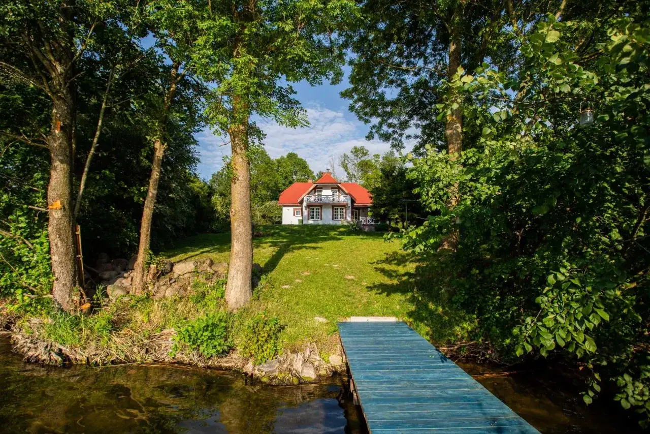 Ustronny domek nad jeziorem, otoczony zielenią, z niebieskim pomostem prowadzącym do wody. Idealne miejsce na wypoczynek.