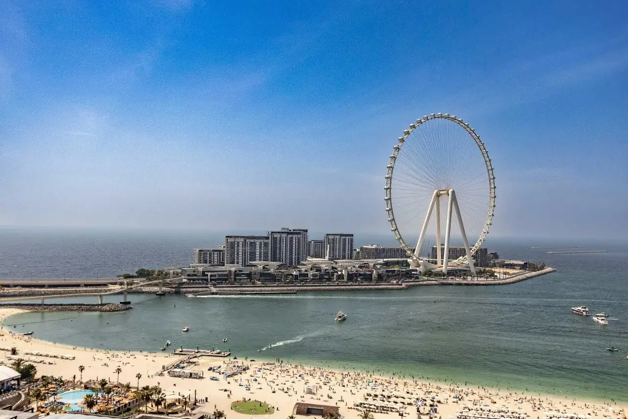 Wyspa z diabelskim młynem w Dubaju, plaża pełna ludzi i błękitne morze pod bezchmurnym niebem.