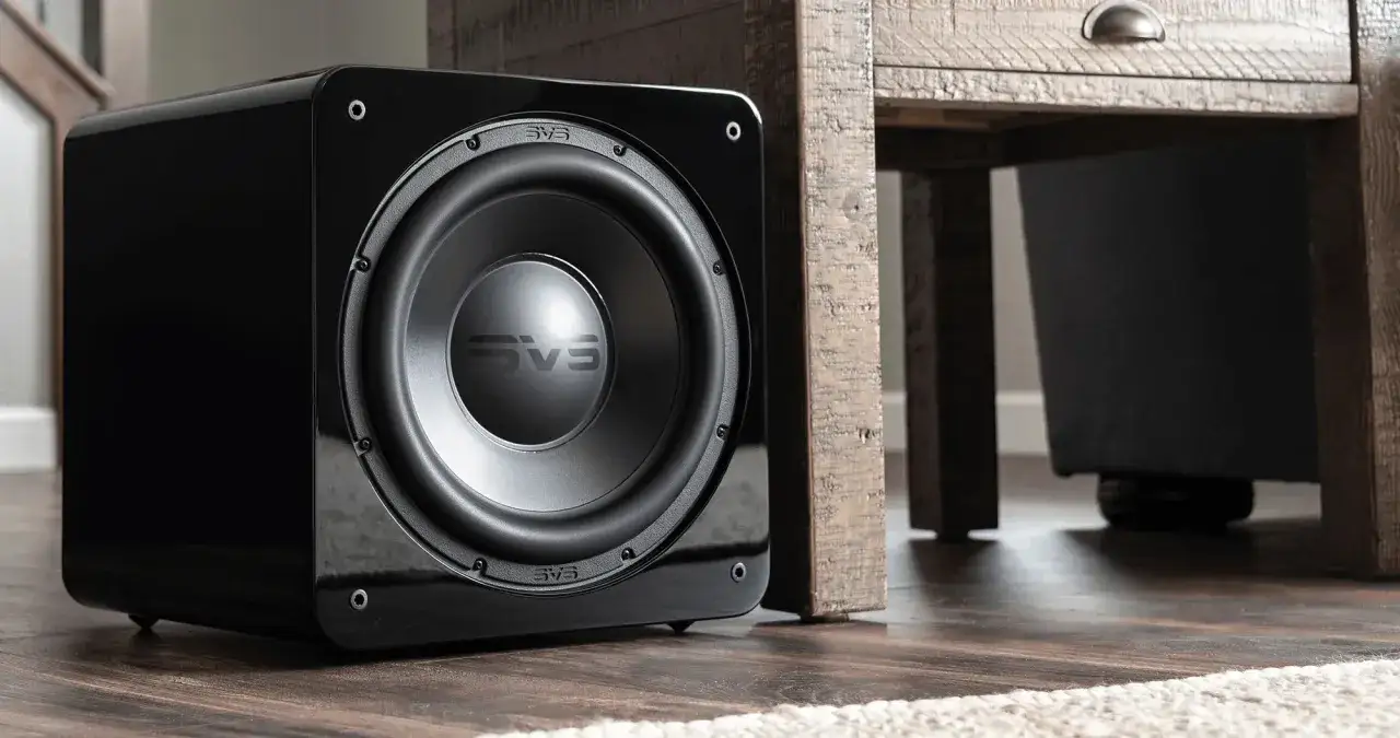Un caisson de basse noir brillant SVS est plac&eacute; pr&egrave;s d'un meuble en bois rustique, optimisant le placement caisson de basse pour un son immersif.