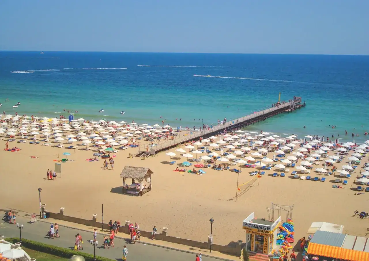 Słoneczny brzeg pogoda dopisuje. Tłumy plażowiczów, białe parasole i molo w turkusowym morzu.