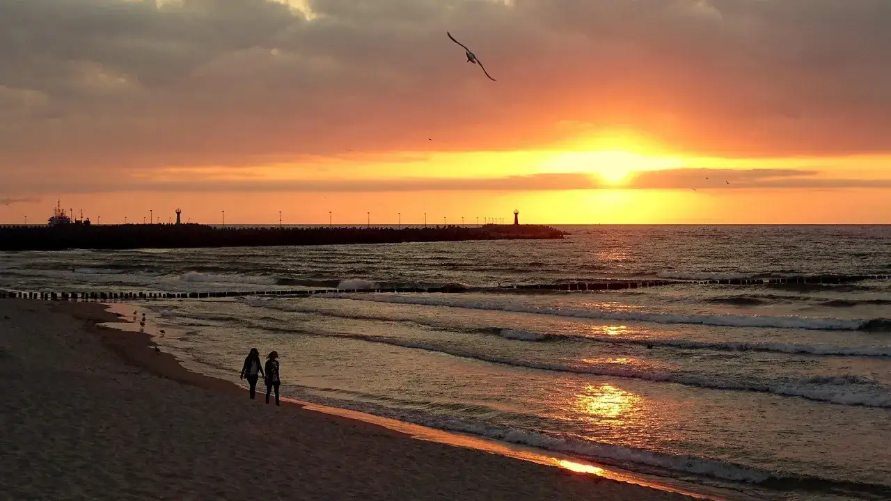 Zachód słońca nad morzem, idealne miejsce, gdzie jechać nad morze w listopadzie. Dwie osoby spacerują po plaży, mewa leci nad falami.