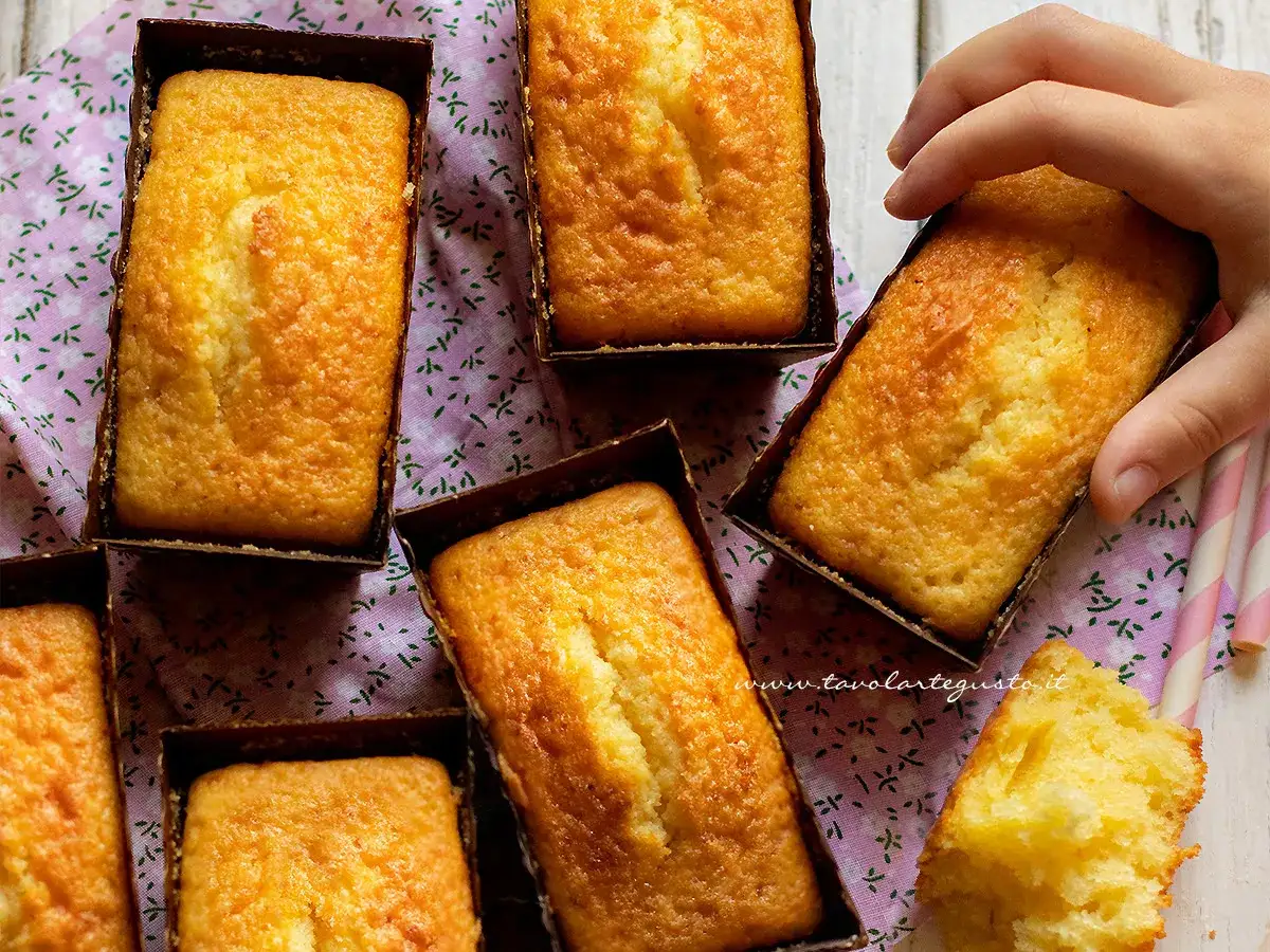 Plumcake yogurt dorati e soffici, pronti per essere gustati. Una mano ne afferra uno, mentre un pezzetto mostra la morbidezza interna.