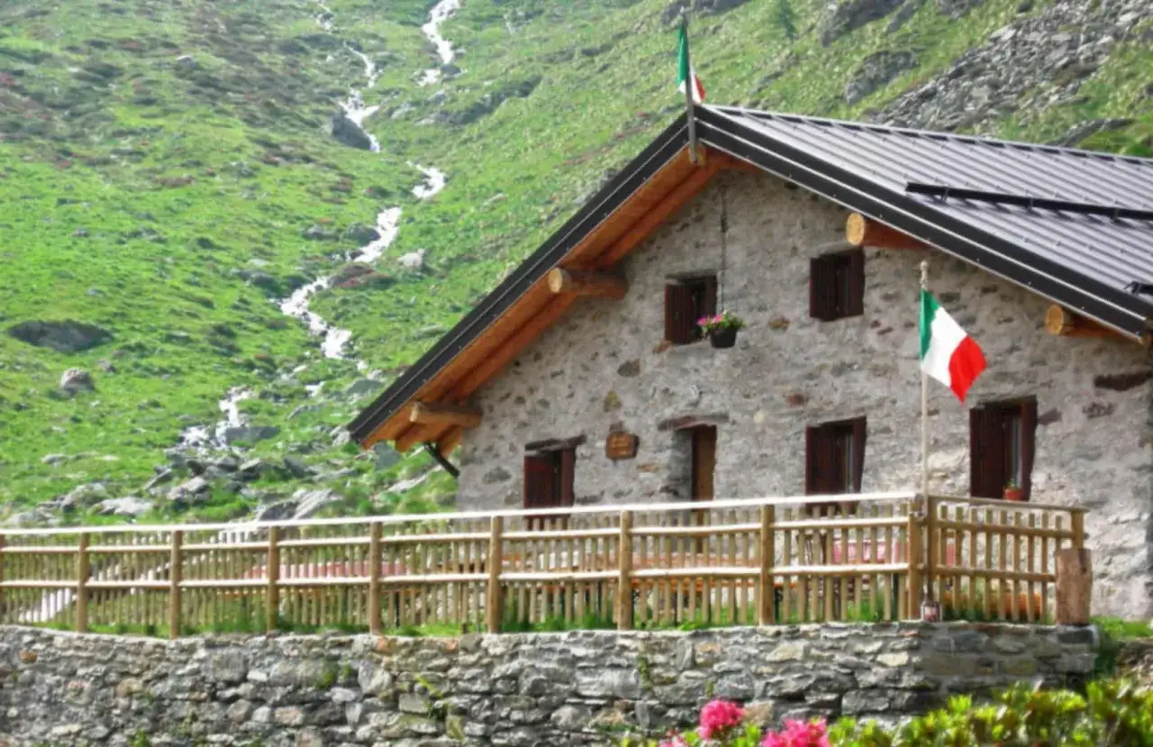 Rifugio in pietra con bandiera italiana, ideale per passeggiate in montagna in Lombardia. Un ruscello scende lungo il pendio verdeggiante.