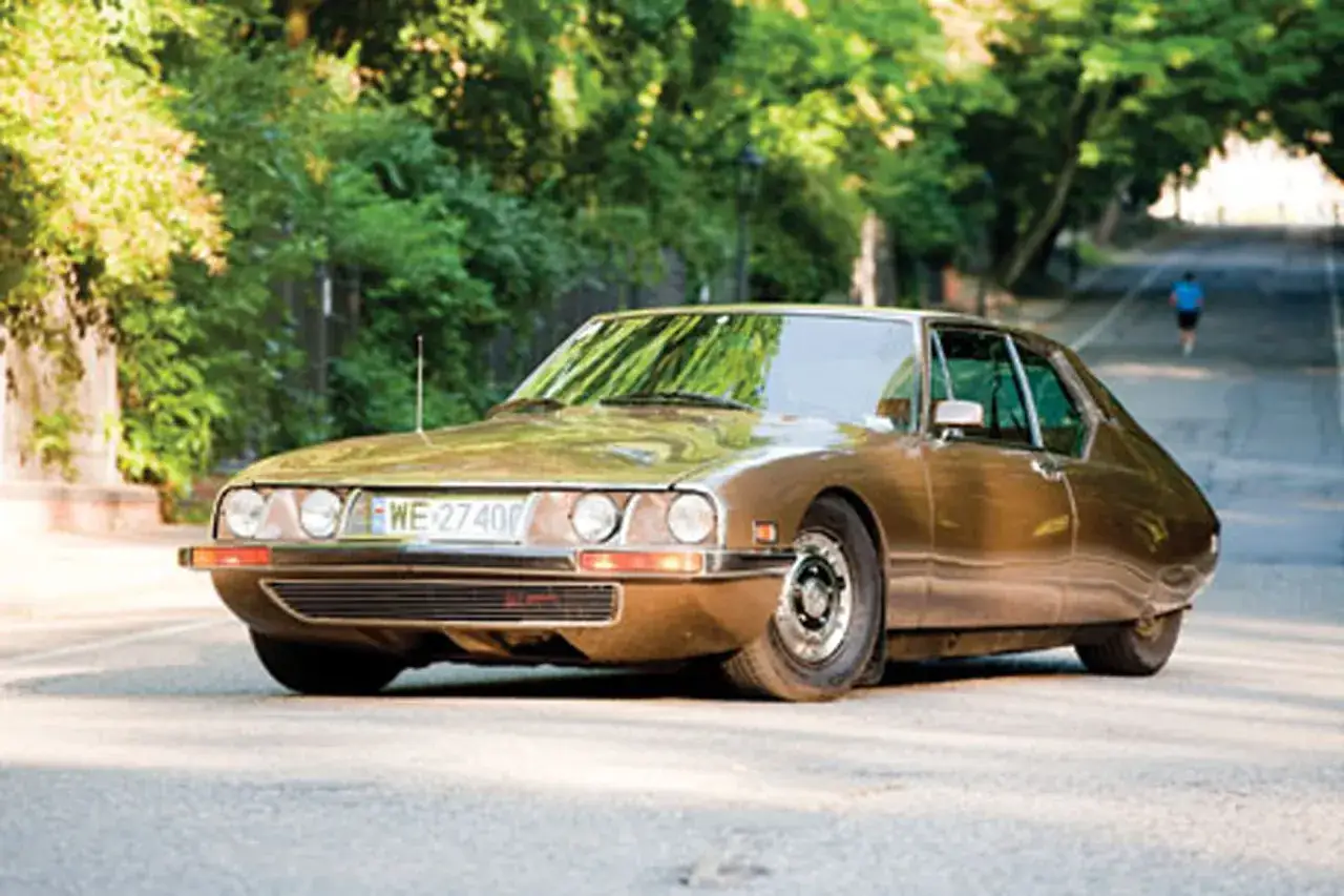 Złoty Citroën SM z lat 70. sunie po asfaltowej drodze, otoczony zielenią.
