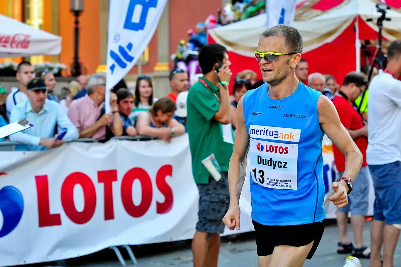 Biegacz z numerem 13, Dudycz, w niebieskiej koszulce, biegnie w maratonie. Widzowie kibicują mu za barierką z napisem LOTOS.
